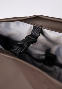 Bruine waterdichte tasinterieur met een zwarte verstelbare riem en plastic gesp detail. Gladde textuur met stiksels langs de randen.