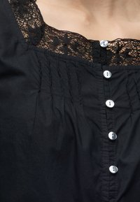 Gros plan d'un chemisier noir avec un bord en dentelle au niveau de l'encolure et une rangée verticale de boutons blancs à l'avant.