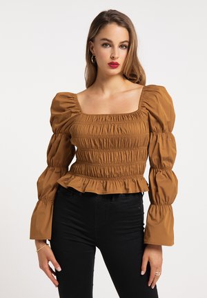 Jonge vrouw met lang haar, dragend een bruine geplooide blouse met pofmouwen en een zwarte broek, staand tegen een effen lichte achtergrond.