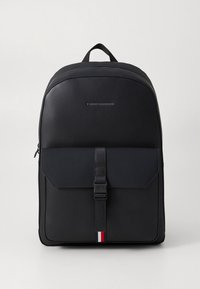 Zaino nero realizzato in materiale sintetico liscio, con chiusura a patta con cinturino, parte superiore arrotondata e un piccolo dettaglio rosso, bianco e blu.