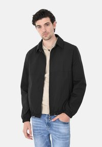Giacca leggera nera con colletto, polsini elasticizzati e vita elasticizzata; abbinata a una camicia beige e jeans blu.
