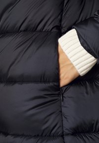 Sort, puffy jakke med et skinnende nylon ydermateriale. Har en sidelomme og en ribbet cremefarvet manchet, der stikker frem fra ærmet.
