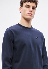 Tmavomodrá mikina z mäkkej látky, so zaokrúhleným výstrihom a malým, okrúhlym logom na hrudi. Dlhé rukávy s rebrovanými manžetami.