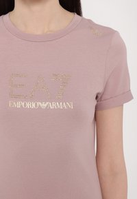 Svetlo ružové tričko s krátkym rukávom. Má zlatý kryštálový logotyp "EA7 EMPORIO ARMANI" a dekoratívne detaily na pleci. Mäkká tkanina.