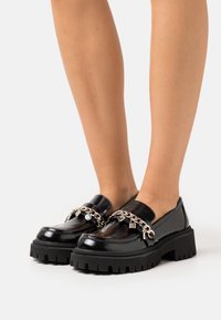 Mocassins en vernis noir avec des semelles en caoutchouc épaisses, accents en chaîne argentée ornés de charme. Fini lisse et design à bout rond.