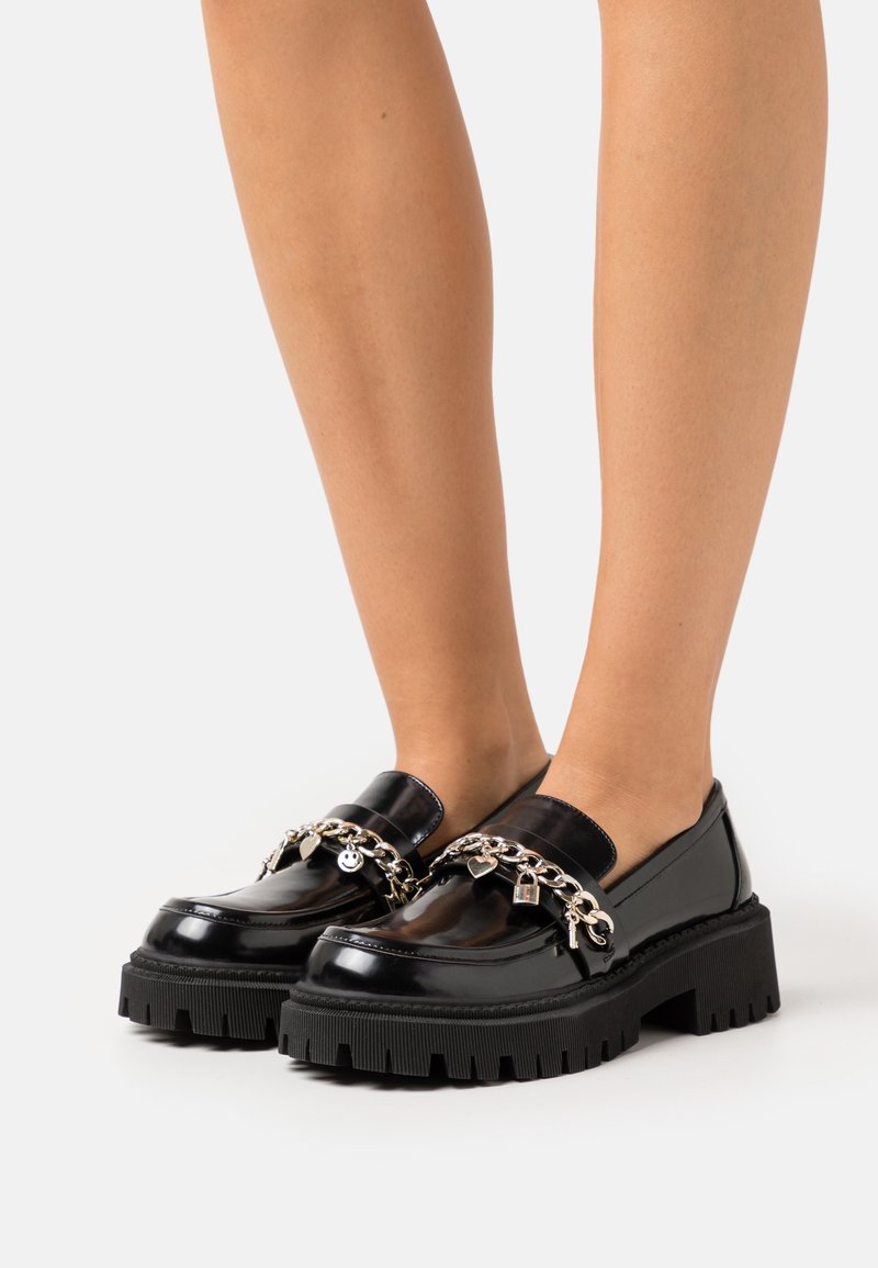 Mocassins en vernis noir avec des semelles en caoutchouc épaisses, accents en chaîne argentée ornés de charme. Fini lisse et design à bout rond.