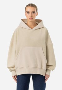 Felpa oversize beige realizzata in tessuto morbido, con tasca frontale, maniche cuffiate e cappuccio con cordino. Dettagli ricamati sottili.