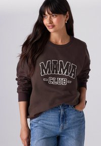 Brūna sporta krekls ar baltiem, treknrakstā uzrakstiem "MAMA CLUB". Garās piedurknes ar ribbinātajām aprocēm. Kombinēts ar ziliem, brīvi pieguļošiem džinsiem.