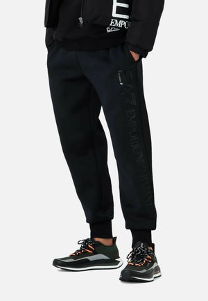Pantaloni sportivi neri a vestibilità slim, con un logo in finta pelle sul lato, abbinati a sneaker sportive nere e verdi con dettagli arancioni.