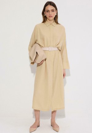 Žena v béžových midi šatách so skladacím opaskom držiaca zodpovedajúci kabát, v kombinácii s béžovými špicatými balerínami so zadným popruhom, stojaca na bielom pozadí.