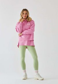 Różowa, oversized bluza z kapturem, z kieszenią z przodu i logo, nałożona na krótszą bluzkę. Zielone legginsy i jasne sneakersy z teksturowaną podeszwą.