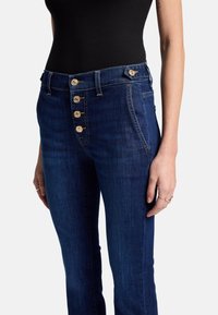 Jeans in denim blu scuro con vita alta, dotati di cinque bottoni dorati e dettagli di cucitura sottili sulle tasche anteriori.
