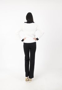 Pull blanc à côtes avec un col noir volanté, associé à un pantalon noir droit et des chaussures à talons beiges. Fond uni.