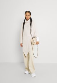 Poncho beige con colletto alto, caratterizzato da un orlo inclinato. Indossato sopra pantaloni larghi beige e abbinato a sneaker bianche e una borsa piccola.