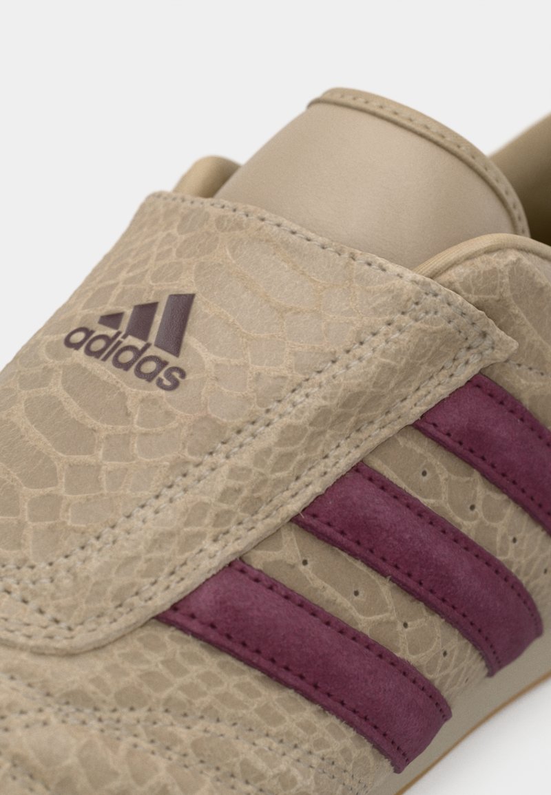 Baskets beige avec une finition texturée rappelant une peau de reptile, arborant des rayures bordeaux. Le design comprend un logo adidas sur le côté.