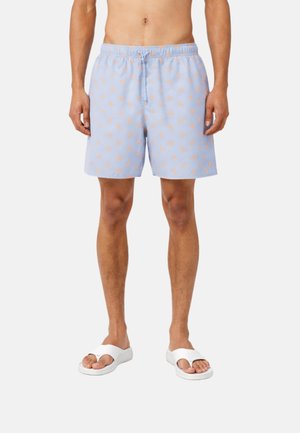 Hombre con pantalones cortos de baño azul claro con pequeños patrones naranjas y sandalias blancas tipo slide, de pie contra un fondo liso.