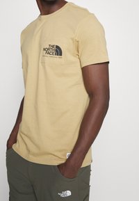 Muž v béžovém tričku s krátkým rukávem s logem "The North Face" na kapse a tmavě zelených kalhotách s logem na stehně, ruce v kapsách.