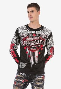 Langärmliges schwarzes Hemd mit roten und weißen grafischen Designs, einschließlich Logos und Mustern. Kombiniert mit abgetragenen, bemalten Jeans.