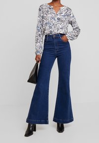 Jeans en denim bleu foncé évasés portés avec une chemise à manches longues blanche et bleue à motifs, accompagnés d'un sac à main noir. Bottes à talons noires.