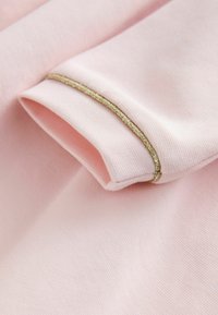 Vaaleanpunainen kangas, jossa on teksturoitu pinta, ja kultainen metallinen reunus käänteellä sekä sileät reunat.