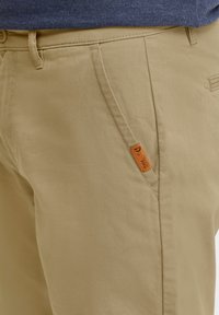 Beige katoenen broek met een gladde textuur, voorzien van zijzakken, een gestikte afwerking en een klein oranje label vlakbij de tailleband.