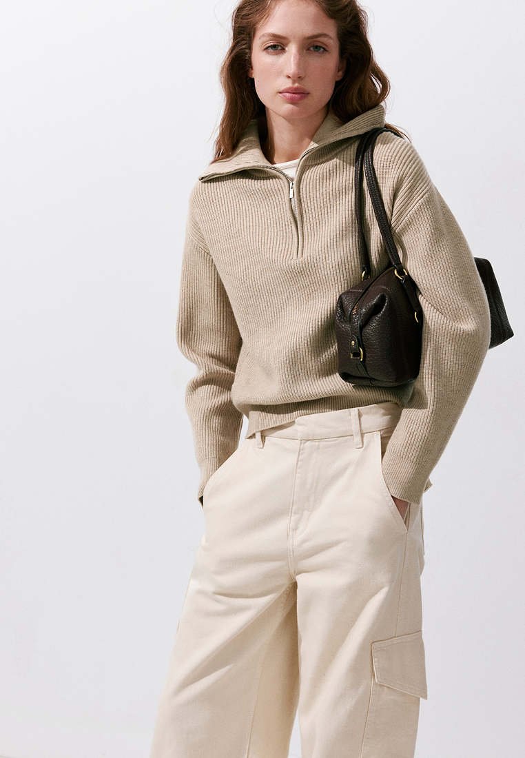 Femme portant un pull beige côtelé zippé et un pantalon large crème, portant un sac à bandoulière en cuir noir, debout sur fond blanc.