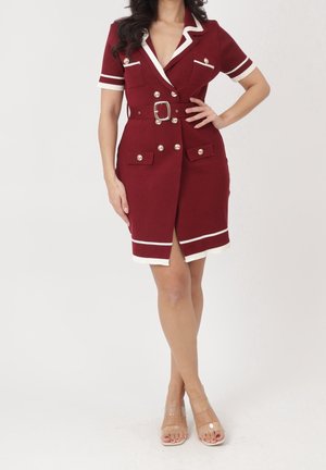 Frau trägt ein rotes Blazer-Kleid mit Gürtel, weißem Besatz und goldenen Knöpfen, kombiniert mit transparenten, offenen High Heels, steht vor weißem Hintergrund.