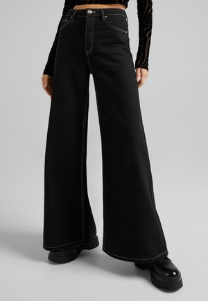 Jeans anchos negros con costuras blancas en contraste, de cintura alta y cierre de botón. Estilizados con botas de plataforma negras y gruesas.