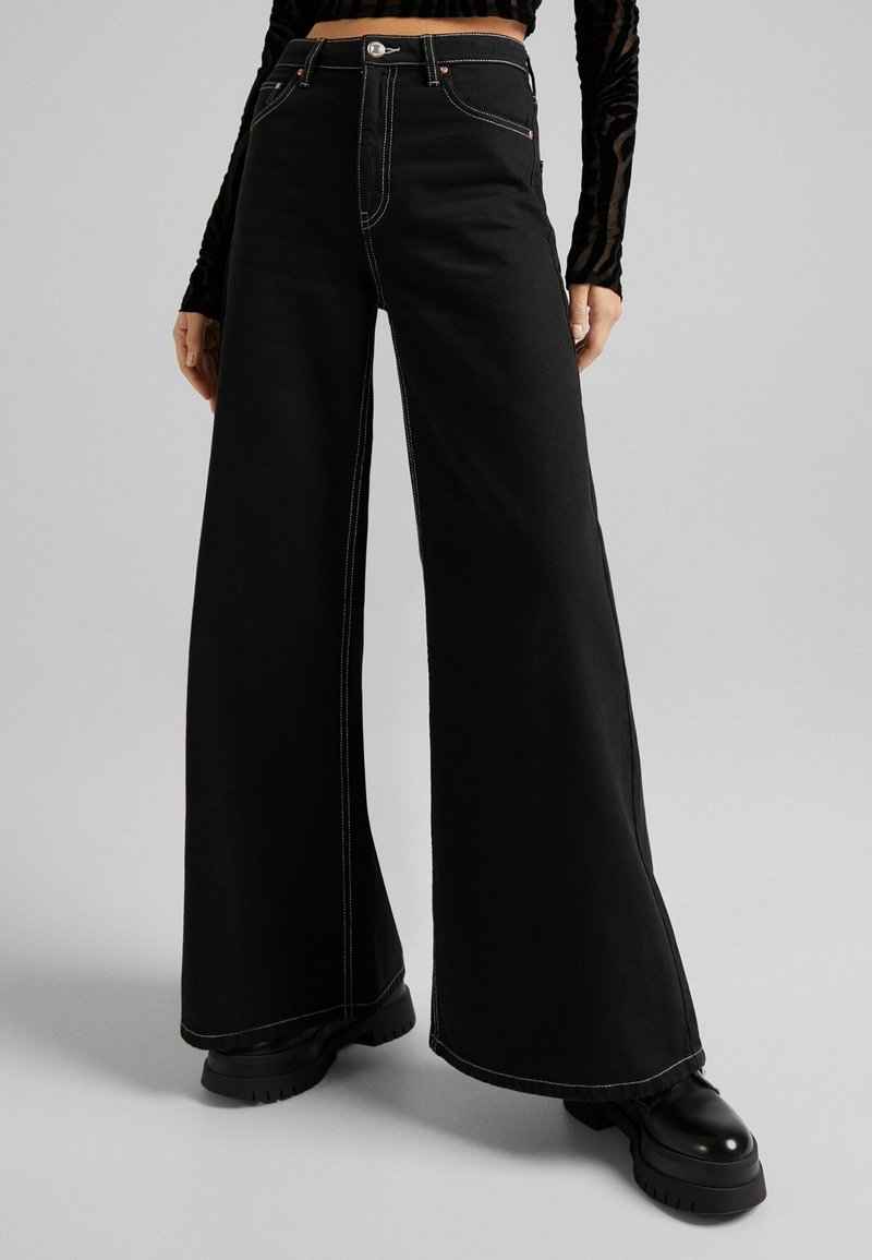 Svart vida jeans med kontrasterande vit sömnad, hög midja och knappstängning. Stylandes med kraftiga svarta plattformsskor.