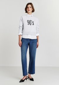 Felpa grigia con la stampa "90's" sul davanti, indossata sopra una camicia bianca con colletto, abbinata a jeans larghi blu e mule neri con tacco.
