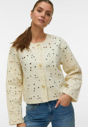 Nő hosszú barna hajjal lófarokban, krémszínű, kivágott gombos dzsekit és kék farmert visel, egyik kezét zsebében tartva áll.