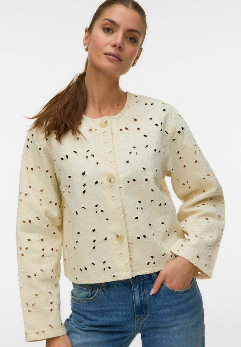 Femme aux longs cheveux bruns attachés en queue de cheval, portant une veste crème à œillets boutonnée et un jean bleu, debout avec une main dans la poche.