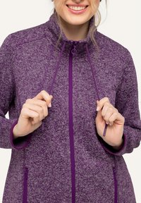 Giacca in pile viola con un motivo texturizzato, colletto alto, chiusura a zip completa e dettagli con coulisse. Presenta tasche con zip sui lati.