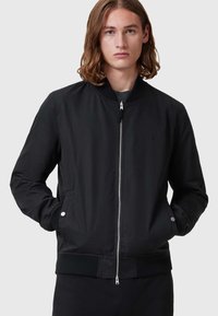 Schwarze Bomberjacke mit einem Reißverschluss vorne, gerippten Bündchen und Kragen, mit Seitentaschen und einem dezenten Logo auf der Brust. Glatte Stoffoberfläche.