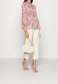 Femme portant un pantalon cropped beige, un chemisier à motifs rose et beige, des sandales à talons transparents, tenant un sac à main blanc avec une bandoulière en chaîne dorée.