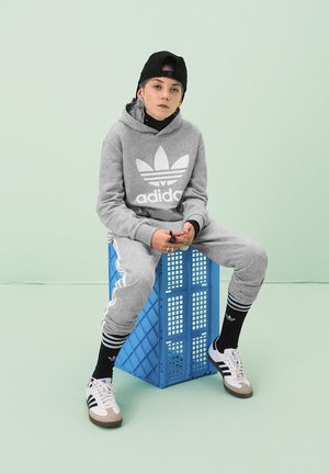 Felpa grigia Adidas e pantaloni della tuta con strisce bianche, calze nere e scarpe Adidas. Seduto su un crate blu contro uno sfondo verde chiaro.