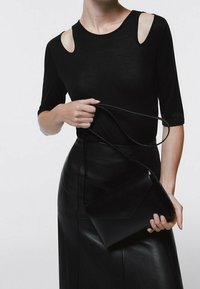 Top aderente nero con ritagli sulle spalle, abbinato a una gonna midi in pelle nera. La modella tiene una piccola borsa a tracolla nera.