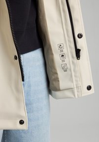 Chaqueta impermeable beige con cierre de cremallera, herrajes negros y costuras selladas térmicamente. Etiquetas de cuidado visibles en el interior de la manga.