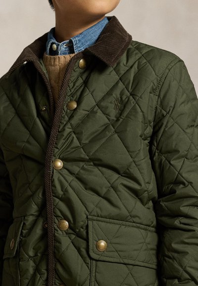 Kind draagt een groene gewatteerde jas met een bruine corduroy kraag, koperen drukknopen, een beige trui en een blauw spijkerbroekhemd eronder.