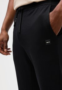 Black jogger pants made from textured fabric with an elastic waistband and drawstring. Features a small logo patch on the pocket.