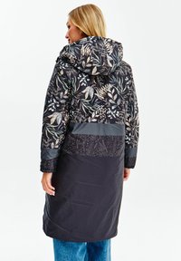 Zateplený kabát se květinovým vzorem v odstínech zelené a krémové na tmavém pozadí, s černou spodní částí a kapucí.