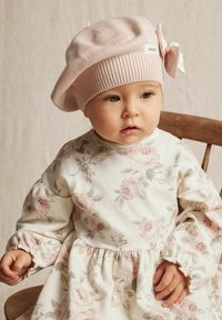 Ljust rosa stickad beret med en rosett, tillsammans med en vit klänning med rosa blommönster och puffärmar. Trästol i bakgrunden.