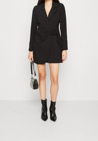 Vestido blazer negro con un detalle de nudo en la cintura, mangas largas y un escote en V profundo. Combinado con botas negras de tobillo y un bolso pequeño.