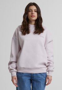 Lys rosa genser med rund hals og ribbestrikkede mansjetter, med en subtil brodert logo på brystet. Brukt med blå jeans.