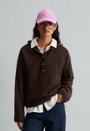 Vrouw draagt een bruin gebreid vest met knopen over een wit overhemd, blauwe spijkerbroek, roze pet en grote bruine bril, staand met één hand in de zak.