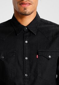 Camisa de mezclilla negra con un cierre de botones en la parte delantera, dos bolsillos en el pecho, botones a presión plateados y un acabado ligeramente texturizado. Etiqueta de acento roja en el bolsillo izquierdo.
