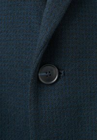 Donkerblauwe gestructureerde stof van een blazer met een enkele ronde zwarte knoop. Close-up weergave die het materiaal en de stiknaden benadrukt.