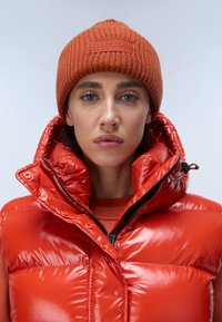 Doudoune rouge sans manches avec col haut, finition brillante et coutures visibles. Associée à un bonnet en tricot orange avec un motif texturé et un logo.