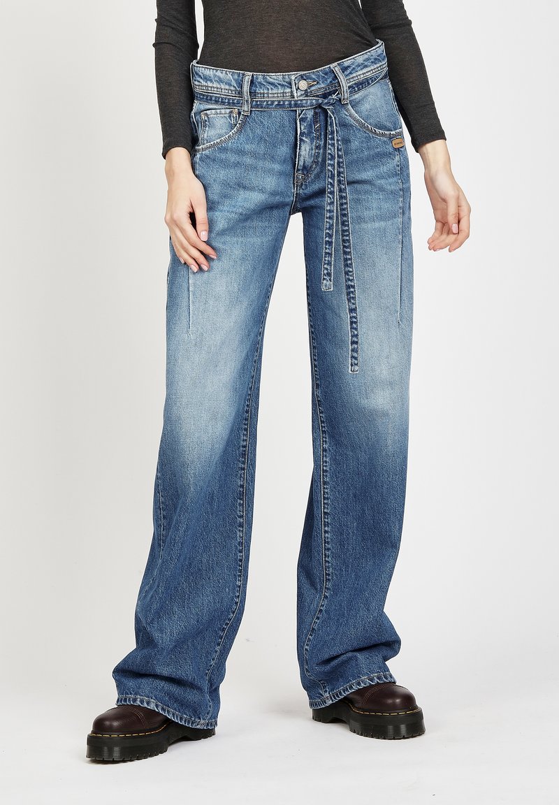 Weite blaue Jeans mit hohem Bund, dekorativem Gürtel und subtilen Verwaschungen. Kontrastnähte und vordere Taschen sind sichtbar.