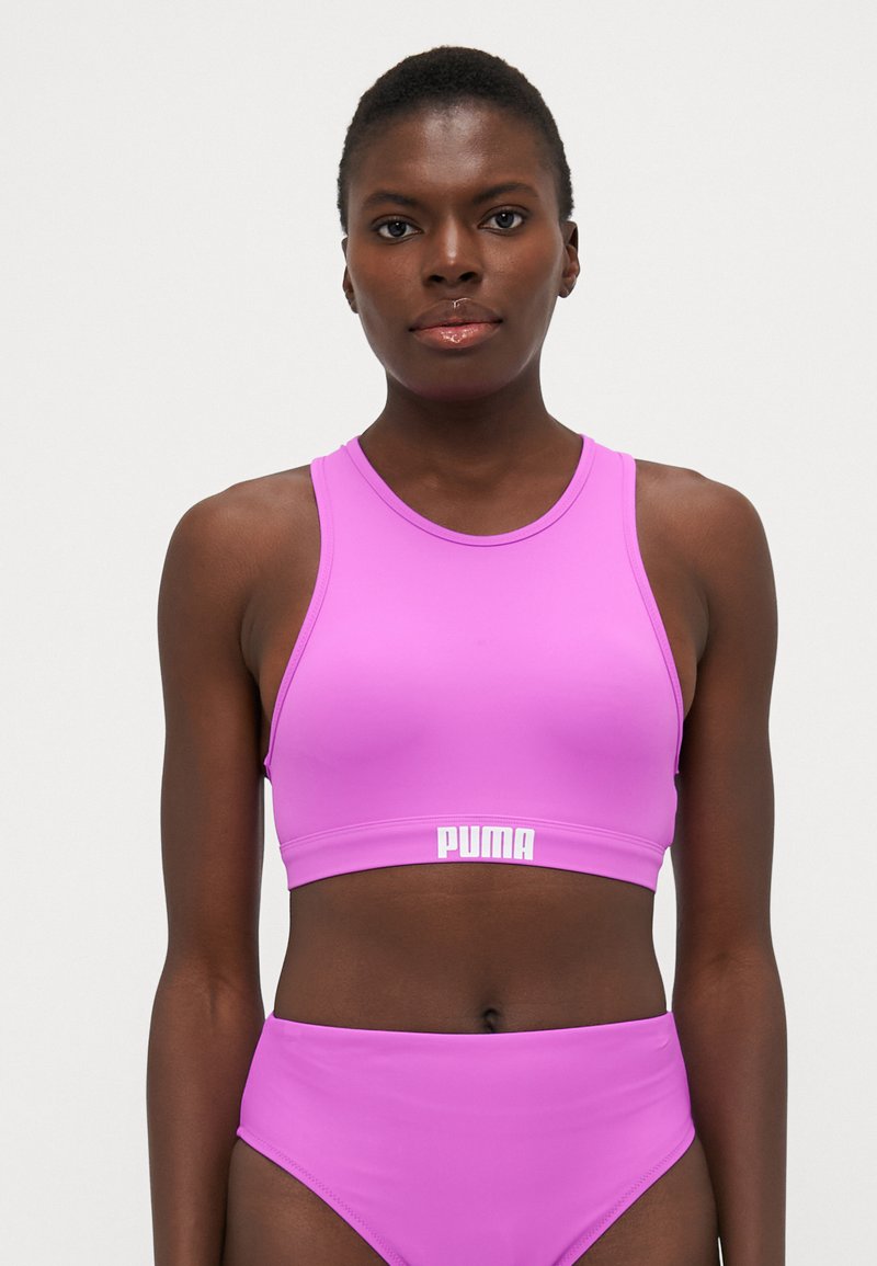Bralette desportivo rosa com decote redondo e design de costas de corredor, apresentando uma faixa de logo branca na parte inferior. Tecido liso e elástico.